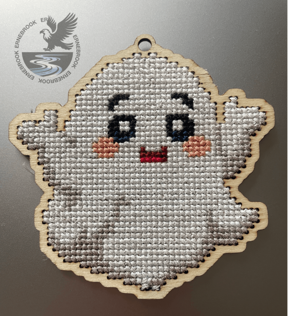 A white friendly ghost cross-stitch home decoration. Cut out of 3mm plywood with a cross-stitch design embroidered on a silver background.