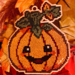 A smiling festive pumpkin cross-stitch home decoration. Cut out of 3mm plywood with a cross-stitch design embroidered on, and resting on Autumn coloured leaves.