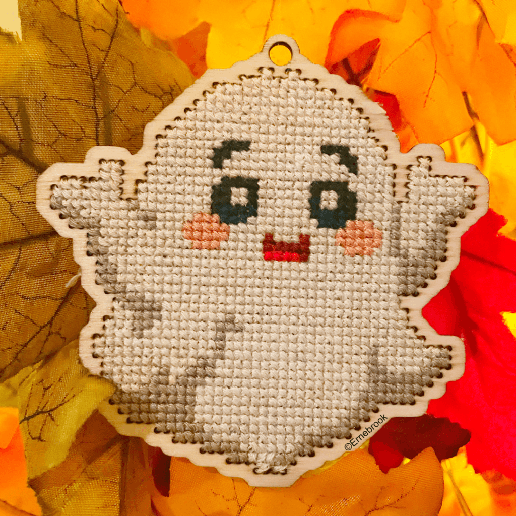 A white, friendly ghost cross-stitch home decoration. Cut out of 3mm plywood with a cross-stitch design embroidered on, and resting on Autumn coloured leaves.