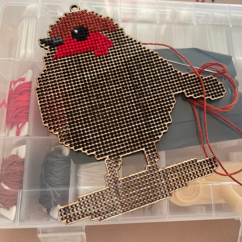 Image of a wooden Robin Redbreast template being completed with cross-stitch and being rested on a box of embroidery threads