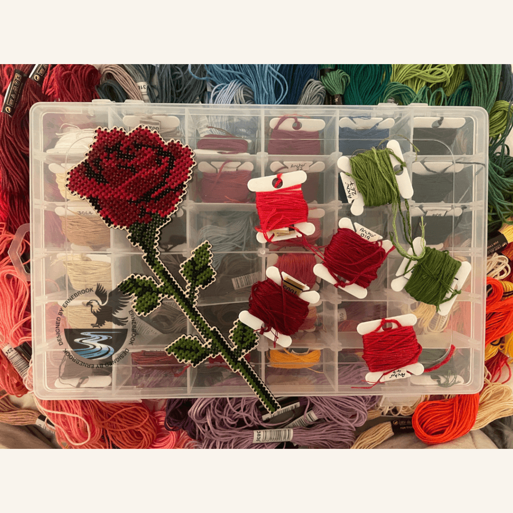 A right-facing single stem rose in cross-stitch with a selection of embroidery threads.