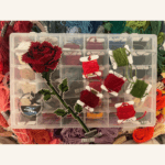 A right-facing single stem rose in cross-stitch with a selection of embroidery threads.