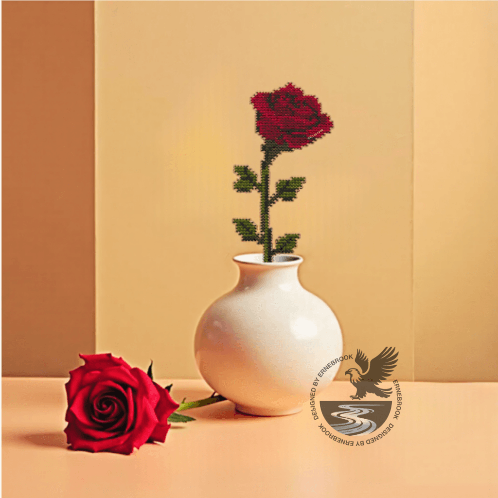 Image of a single red rose template in wood and completed with cross stitch embroidery in a white vase. A real red rose is laying to the side.