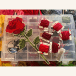 A single straight stem rose in cross-stitch with a selection of embroidery threads.