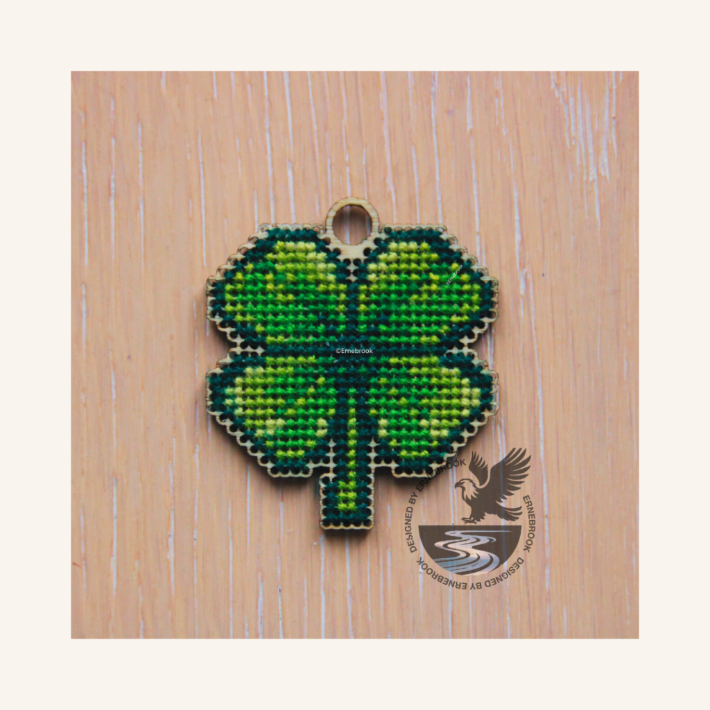 Image of a four-leaf clover cut out of 3mm plywood with a cross-stitch design embroidered, laying on a wooden surface.. The top has a loop incorporated in the wooden template so the finished item could be used as a lucky charm key fob, earring, or pendant.