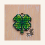 Image of a four-leaf clover cut out of 3mm plywood with a cross-stitch design embroidered, laying on a wooden surface.. The top has a loop incorporated in the wooden template so the finished item could be used as a lucky charm key fob, earring, or pendant.